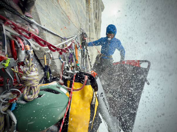 A climber hanging from ropes on a big rock wall, surrounded by climbing equipment, surrounded with a flurry of snow