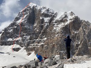 The route in red up a massive, mainly rocky peak. Two climbers observe it with binoculars in front of the mountain.