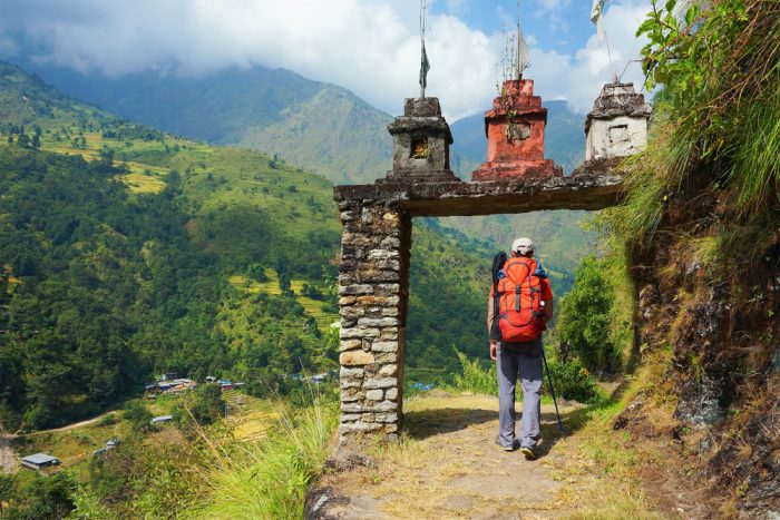A trekker crosses a gate, forest hills and mountains in background.